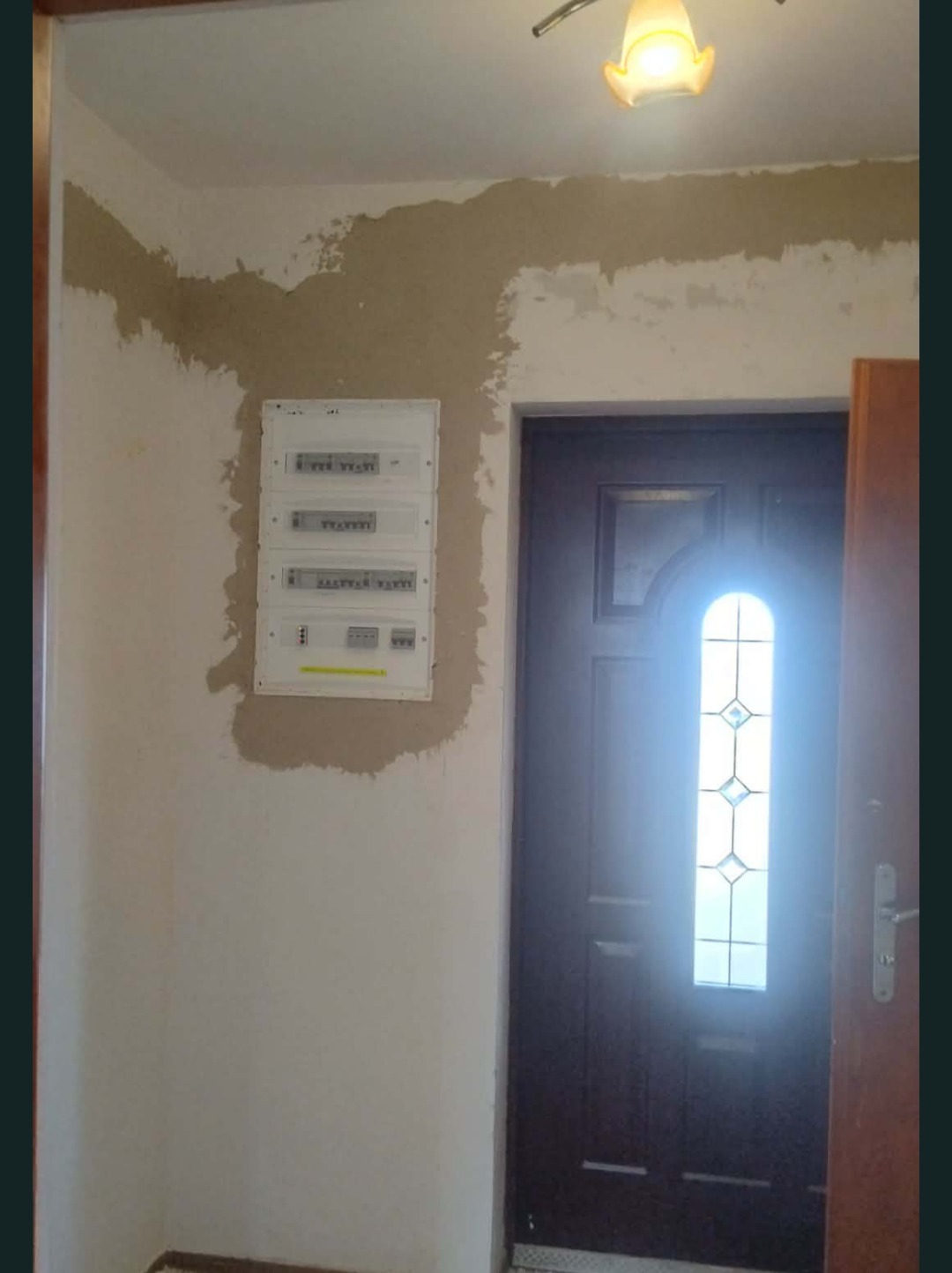 Biała skrzynka elektryczna z bezpiecznikami na ścianie w trakcie montażu, widoczne ślady po szpachlowaniu. W tle ciemne drzwi wejściowe z szybą.