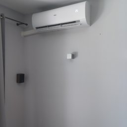 ONE WORLD SOLUTION SPÓŁKA Z OGRANICZONĄ ODPOWIEDZIALNOŚCIĄ - Biała jednostka klimatyzacji Haier typu split zamontowana na szarej ścianie w mieszkaniu. Widoczne korytko maskujące instalację, mały głośnik i karnisz z zasłoną.