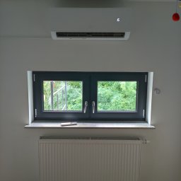 ONE WORLD SOLUTION SPÓŁKA Z OGRANICZONĄ ODPOWIEDZIALNOŚCIĄ - Biała jednostka klimatyzacji typu split zamontowana nad oknem w pokoju. Na parapecie leży pilot. Pod oknem biały grzejnik. Widok na zieleń za oknem.