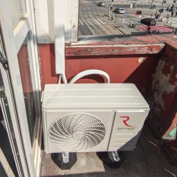 ONE WORLD SOLUTION SPÓŁKA Z OGRANICZONĄ ODPOWIEDZIALNOŚCIĄ - Jednostka zewnętrzna klimatyzacji Rotenso zamontowana na balkonie, widok z góry. Estetyczne podłączenie instalacji, antywibracyjne podstawy. W tle miejski krajobraz.