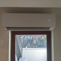 ONE WORLD SOLUTION SPÓŁKA Z OGRANICZONĄ ODPOWIEDZIALNOŚCIĄ - Biała klimatyzacja typu split Haier zamontowana nad oknem w jasnym pokoju. Widoczny okrągły nawiewnik wentylacyjny na ścianie. Na wyświetlaczu klimatyzacji widnieje liczba 18.