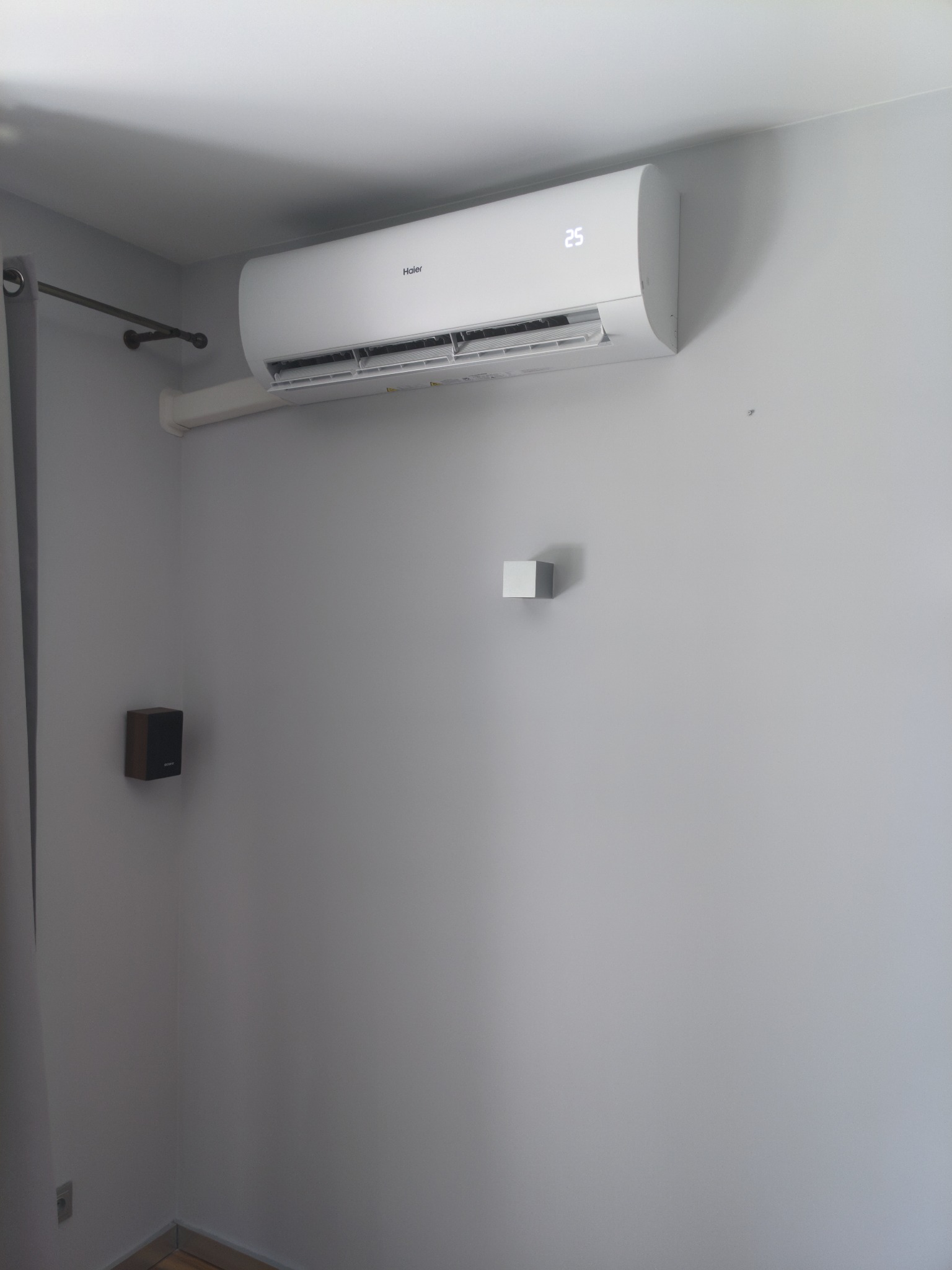 Biała jednostka klimatyzacji Haier typu split zamontowana na szarej ścianie w mieszkaniu. Widoczne korytko maskujące instalację, mały głośnik i karnisz z zasłoną.