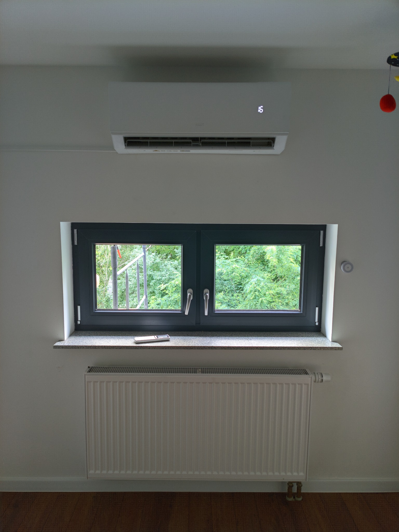 Biała jednostka klimatyzacji typu split zamontowana nad oknem w pokoju. Na parapecie leży pilot. Pod oknem biały grzejnik. Widok na zieleń za oknem.