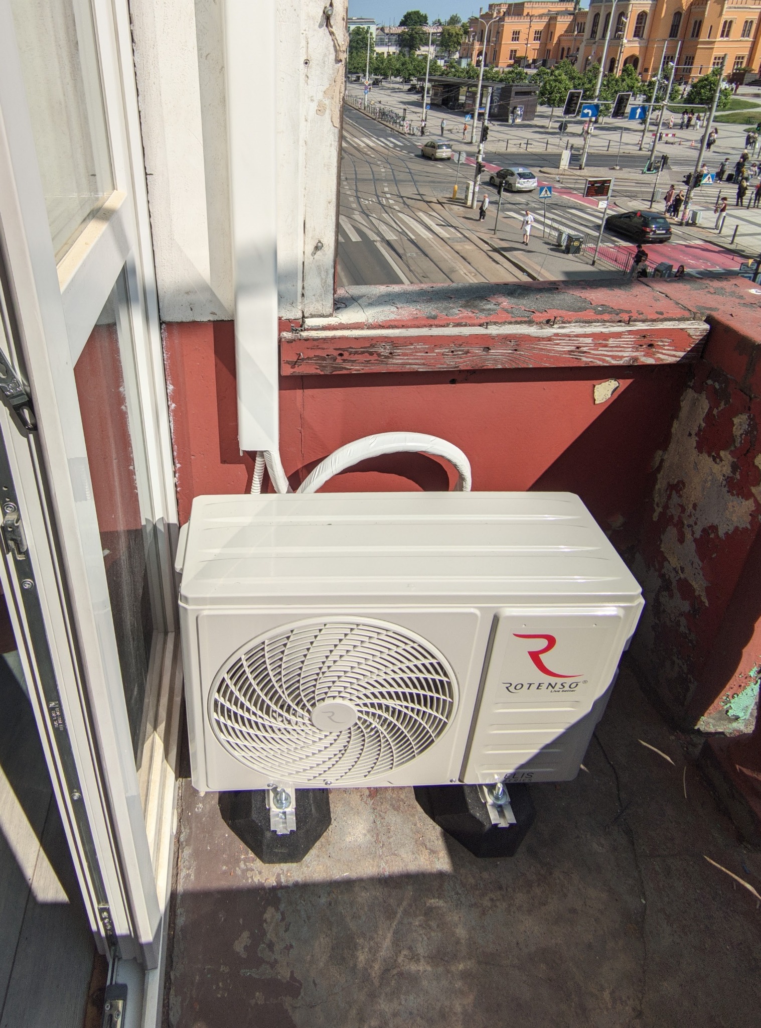 Jednostka zewnętrzna klimatyzacji Rotenso zamontowana na balkonie, widok z góry. Estetyczne podłączenie instalacji, antywibracyjne podstawy. W tle miejski krajobraz.
