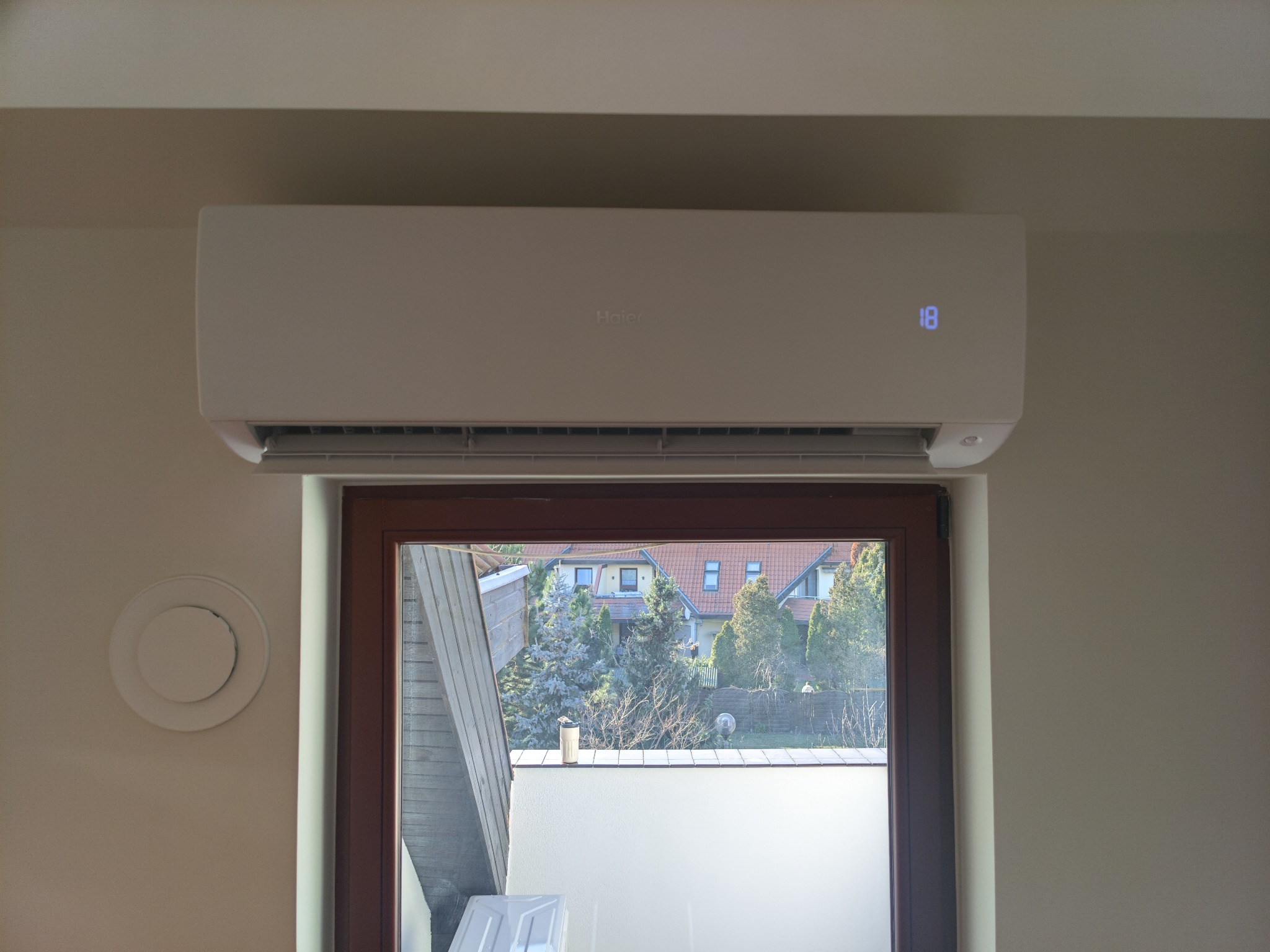 Biała klimatyzacja typu split Haier zamontowana nad oknem w jasnym pokoju. Widoczny okrągły nawiewnik wentylacyjny na ścianie. Na wyświetlaczu klimatyzacji widnieje liczba 18.