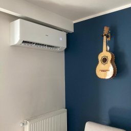 Sklima - Biały klimatyzator Haier na ścianie w pokoju z gitarą meksykańską. Widoczny grzejnik i fragment sofy. Nowoczesne wnętrze z akcentem muzycznym.