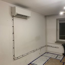 Sklima - Biały klimatyzator na ścianie w trakcie montażu. Widoczne rury instalacyjne prowadzone wzdłuż ściany, zabezpieczona podłoga. Ujęcie z perspektywy narożnika pokoju.