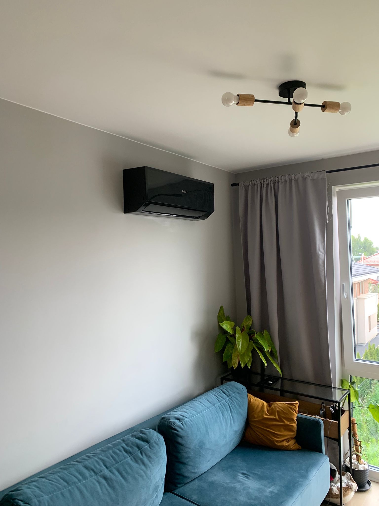 Czarny klimatyzator ścienny Haier w nowoczesnym salonie z turkusową sofą i designerską lampą sufitową. Widok na okno z szarymi zasłonami.
