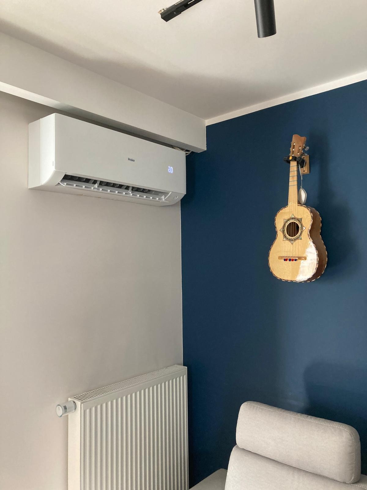 Biały klimatyzator Haier na ścianie w pokoju z gitarą meksykańską. Widoczny grzejnik i fragment sofy. Nowoczesne wnętrze z akcentem muzycznym.