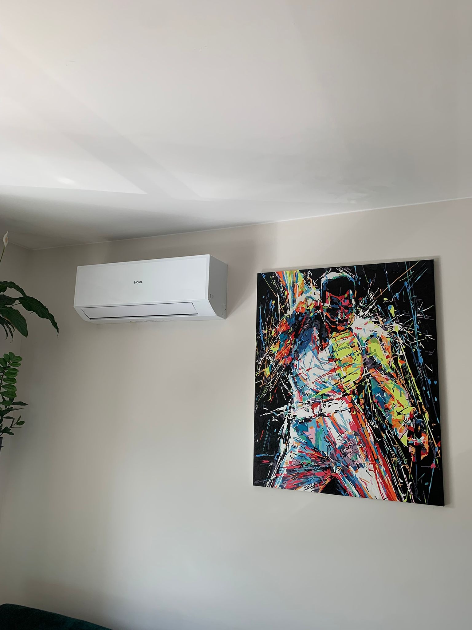 Biały klimatyzator Haier na ścianie w pokoju, obok kolorowy obraz. Widoczny fragment rośliny doniczkowej. Minimalistyczny design wnętrza.