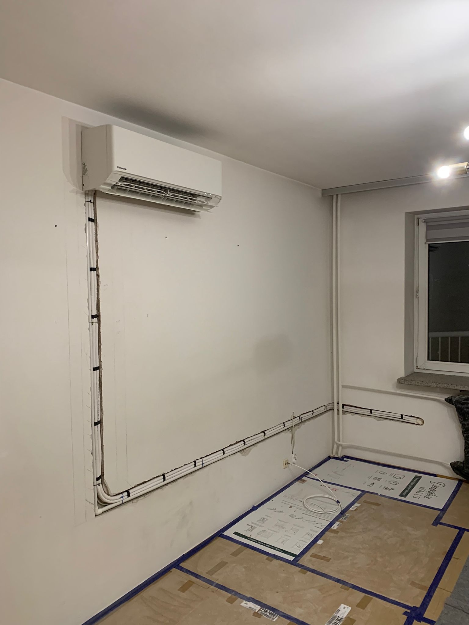 Biały klimatyzator na ścianie w trakcie montażu. Widoczne rury instalacyjne prowadzone wzdłuż ściany, zabezpieczona podłoga. Ujęcie z perspektywy narożnika pokoju.