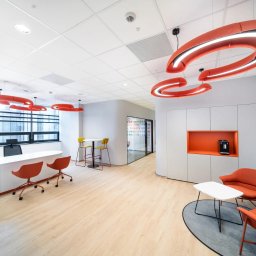 FLASH - Nowoczesne biuro z pomarańczowymi lampami, białymi meblami i drewnianą podłogą. Widoczne krzesła, stolik i zabudowa meblowa z wnęką na ekspres do kawy.