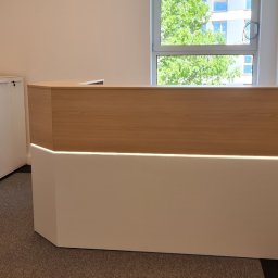FLASH - Nowoczesna lada recepcyjna z oświetleniem LED, wykonana z drewna i białego laminatu, w jasnym biurze z widokiem na zieleń za oknem. Obok szafki biurowe i drukarka.