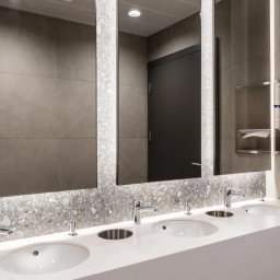 FLASH - Nowoczesna łazienka publiczna z trzema umywalkami w białym blacie, podświetlanymi lustrami i szarymi płytkami. Widoczny dozownik mydła i podajnik ręczników.