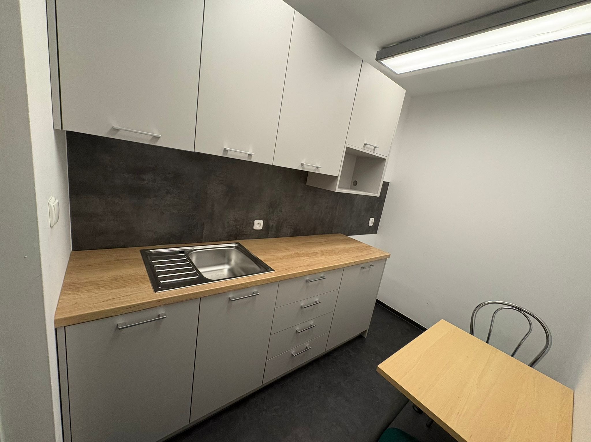 Nowoczesna kuchnia z szarymi szafkami, drewnianym blatem i ciemną ścianą nad zlewem. Widoczny stół z krzesłem. Minimalistyczny design, jasne oświetlenie.