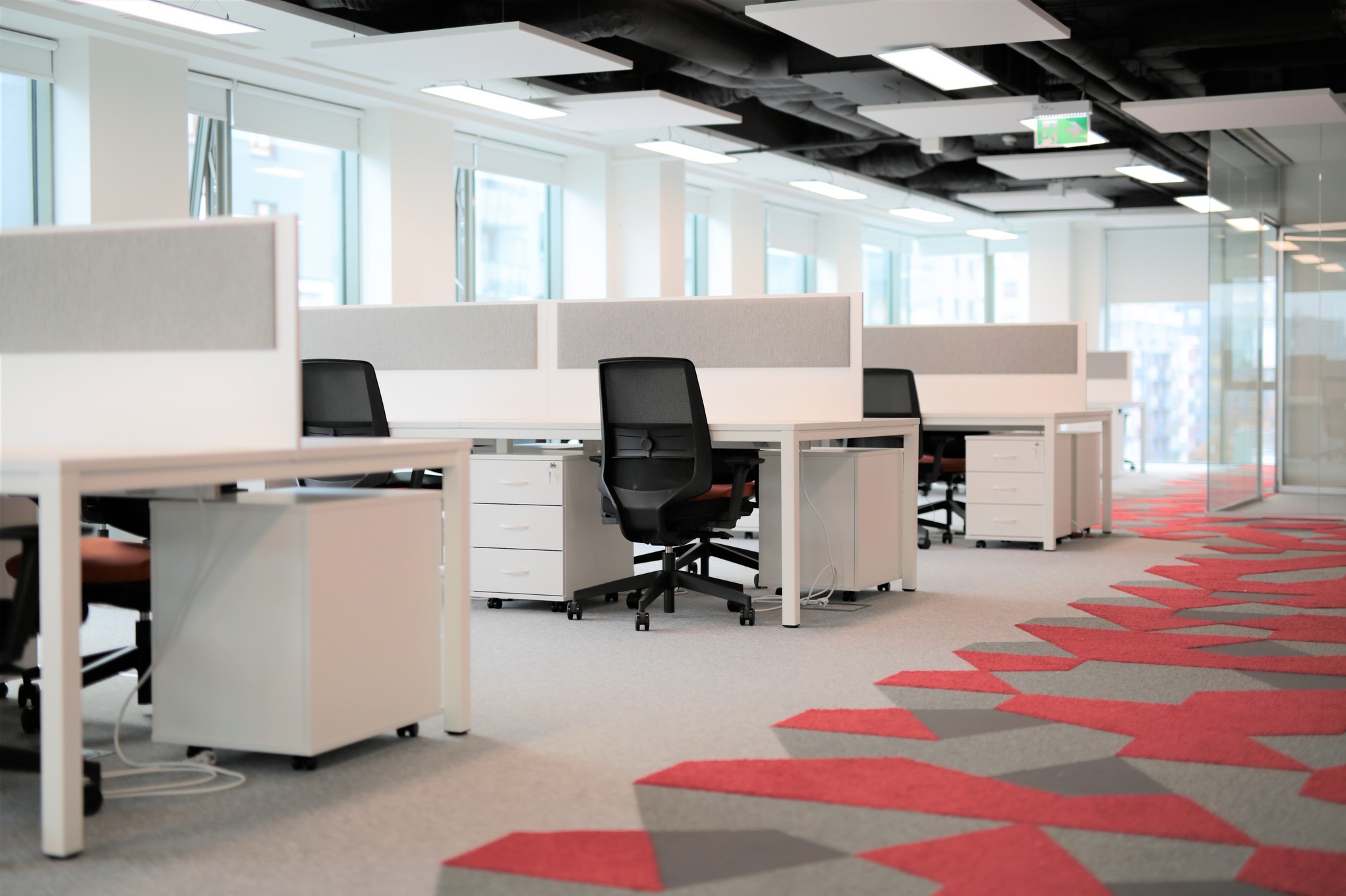 Nowoczesne biuro z białymi biurkami, szarymi przegrodami i wzorzystą wykładziną. Widok na przestrzeń open space z dużymi oknami i naturalnym światłem.