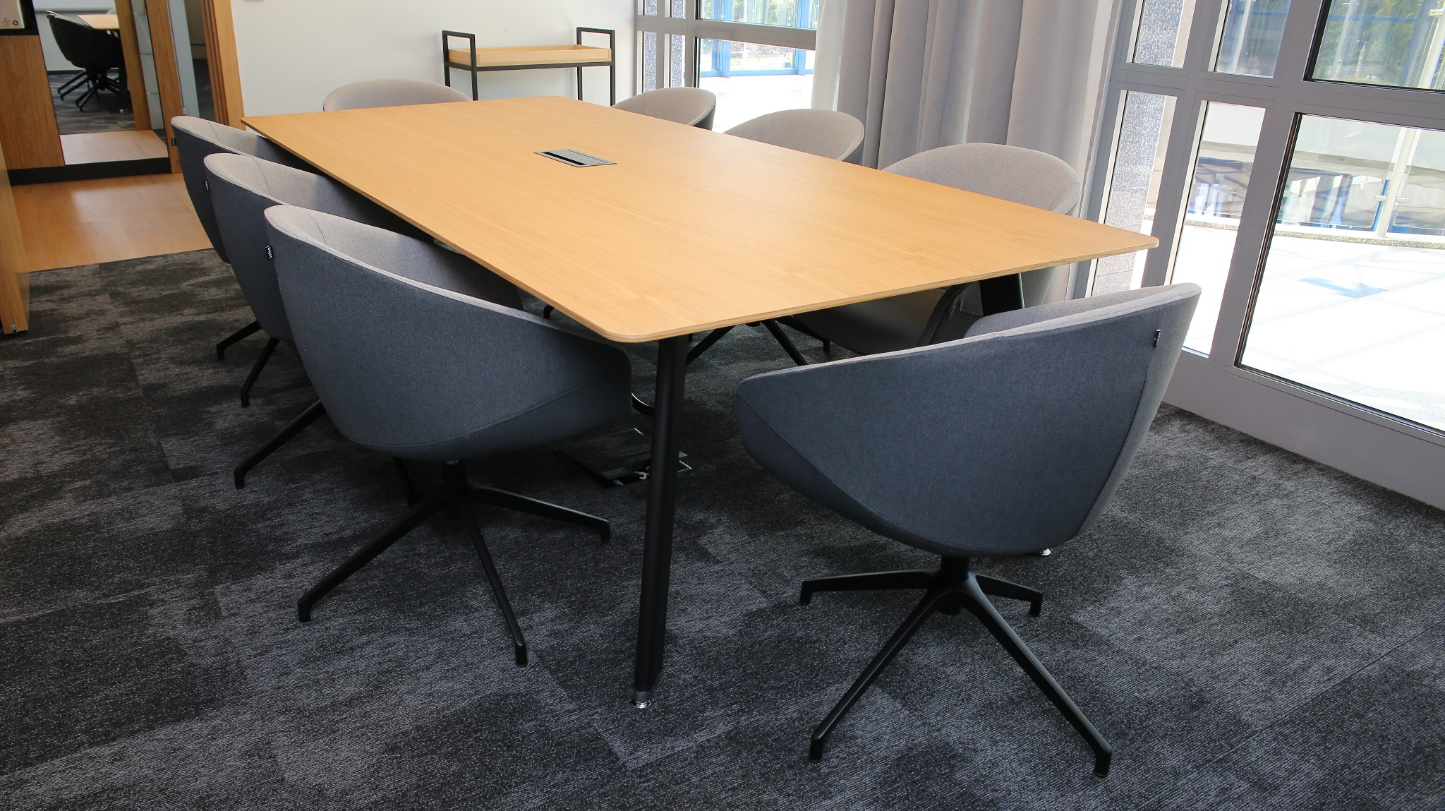 Jasny, drewniany stół konferencyjny z szarymi krzesłami na obrotowych nogach, ustawiony w nowoczesnym biurze z dużymi oknami i szarą wykładziną dywanową.