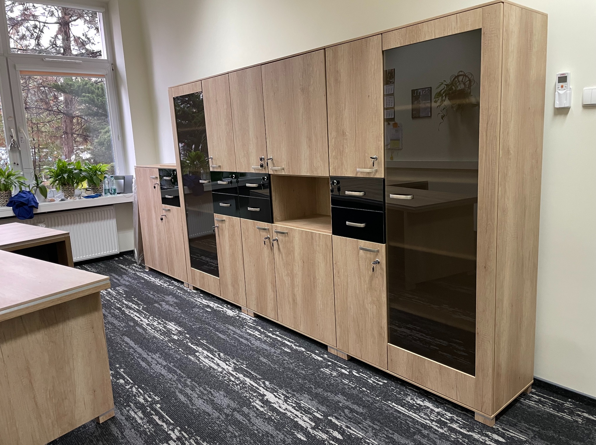 Długa szafa biurowa z jasnego drewna z czarnymi szufladami i przeszklonymi witrynami, widoczna na tle biura z oknem i biurkiem. Nowoczesny design.