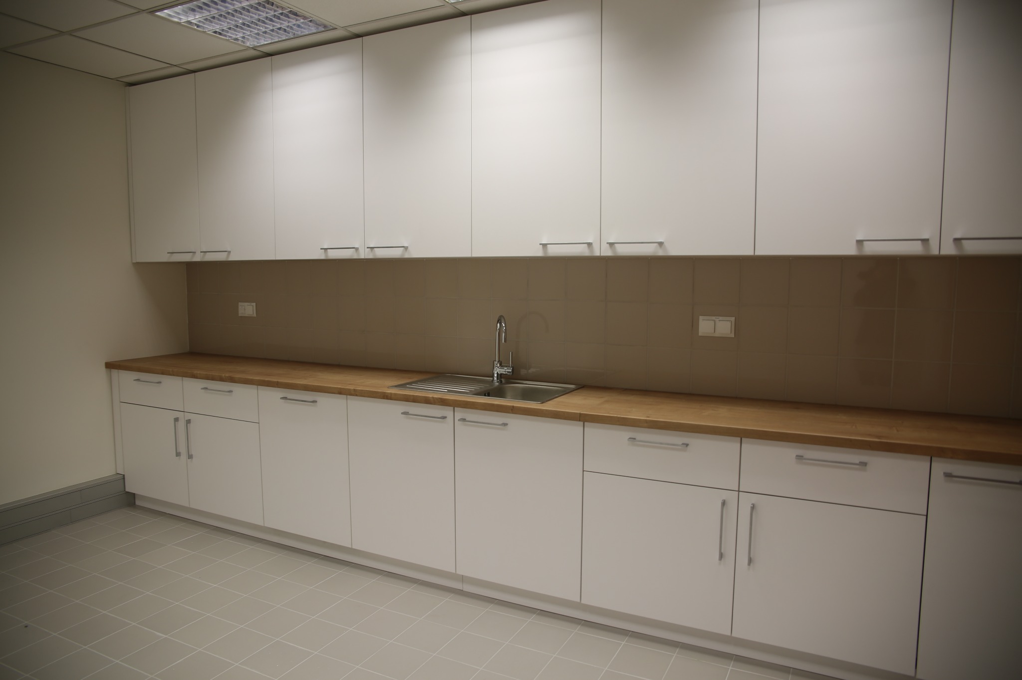 Biała zabudowa kuchenna z drewnianym blatem i beżowymi płytkami. Minimalistyczny design, proste uchwyty, oświetlenie sufitowe. Widok na całą ścianę z szafkami.