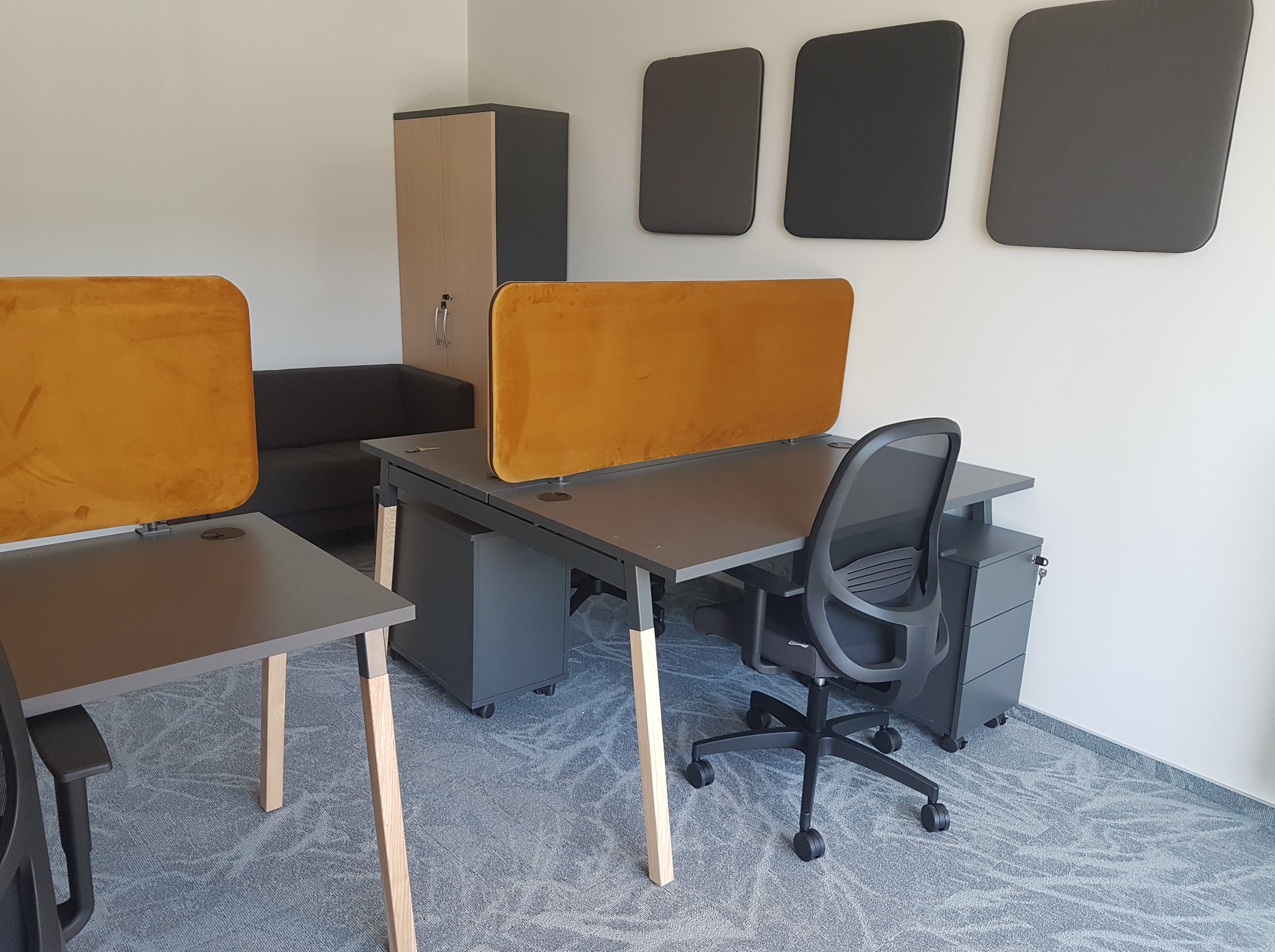 Nowoczesne biuro z szarymi biurkami z żółtymi przegrodami, krzesłem obrotowym i szafką. Na ścianie panele akustyczne. Wykładzina dywanowa w odcieniach szarości.