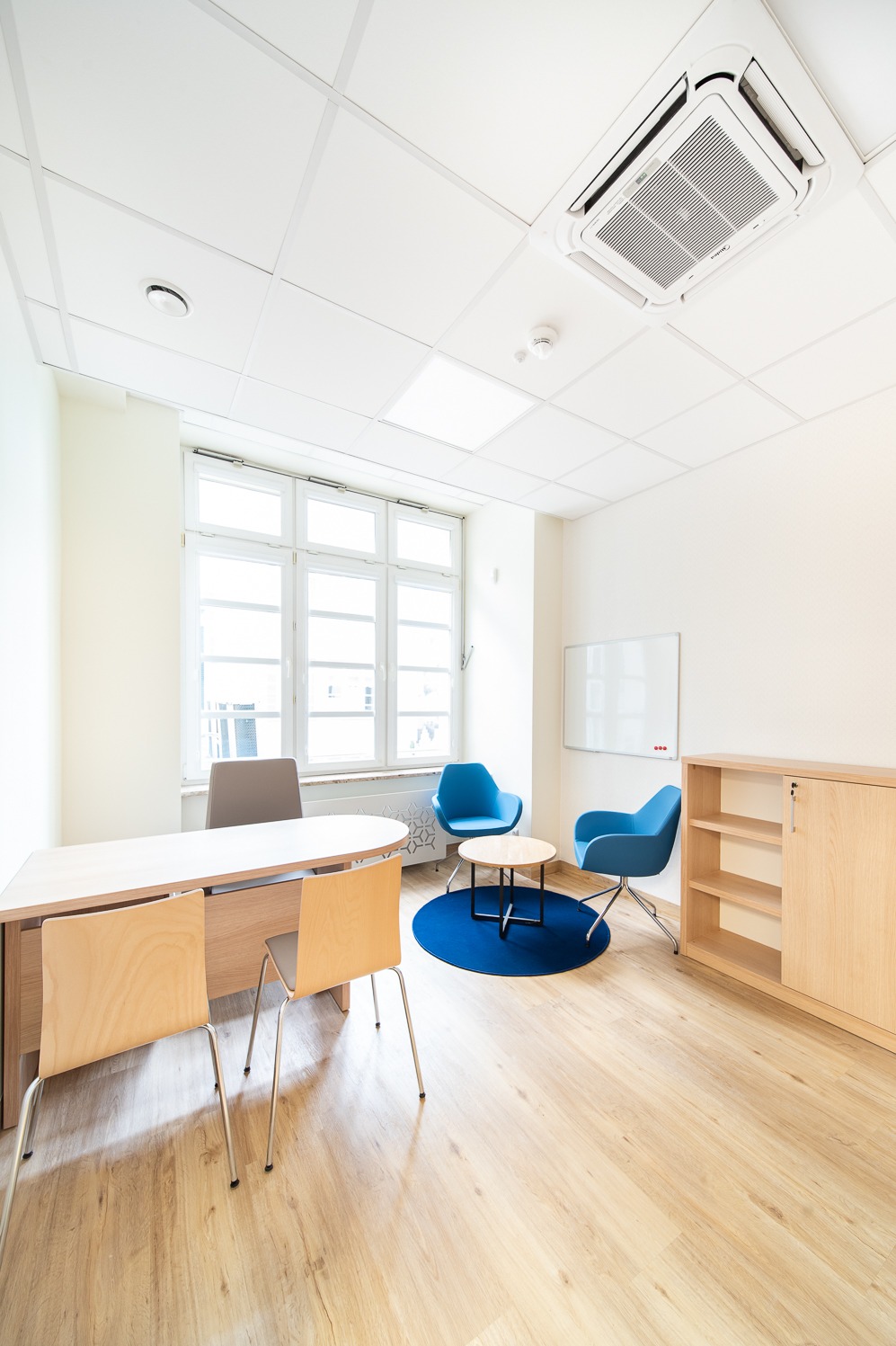 Jasne biuro z drewnianą podłogą, biurkiem, regałem i niebieskimi krzesłami. Białe ściany i sufit z klimatyzacją. Minimalistyczny design, przestronne wnętrze.