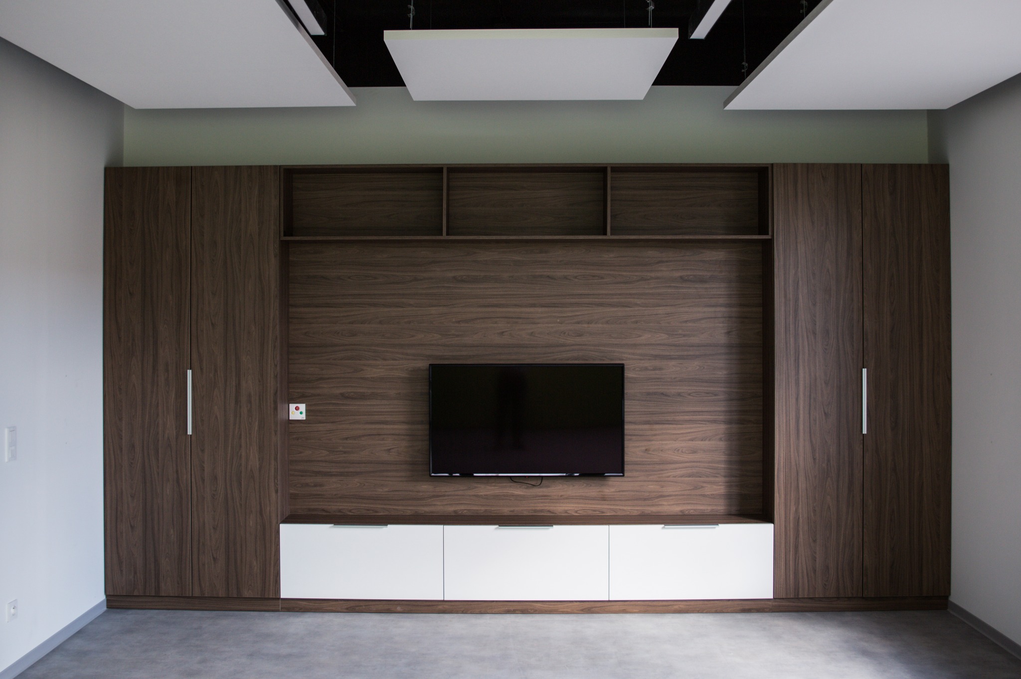 Nowoczesna zabudowa ściany z forniru orzechowego z szafami, półkami i białą szafką RTV pod telewizorem. Minimalistyczne uchwyty, sufit z panelami akustycznymi.