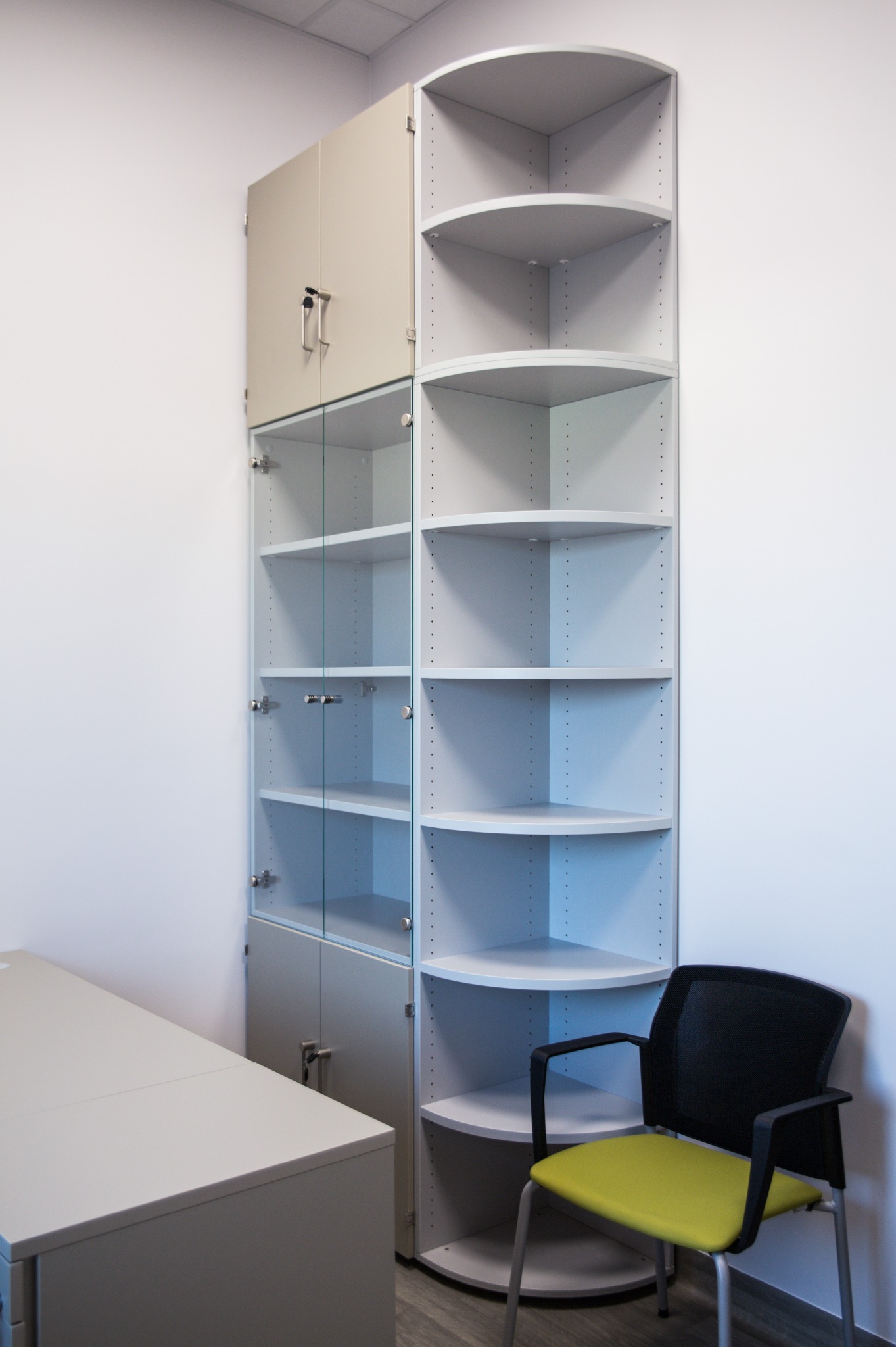 Biało-szara szafa narożna z półkami i przeszklonymi drzwiczkami, uzupełniona biurkiem i krzesłem z żółtym siedziskiem, na tle białej ściany. Minimalistyczny design.