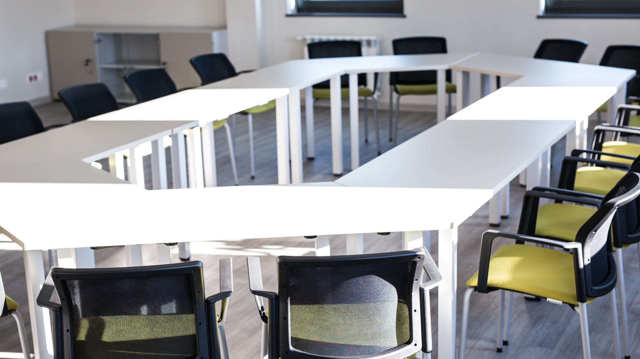 Biały stół konferencyjny w kształcie litery U z krzesłami z siatkowym oparciem i jasnozielonym siedziskiem, oświetlony naturalnym światłem z okna.