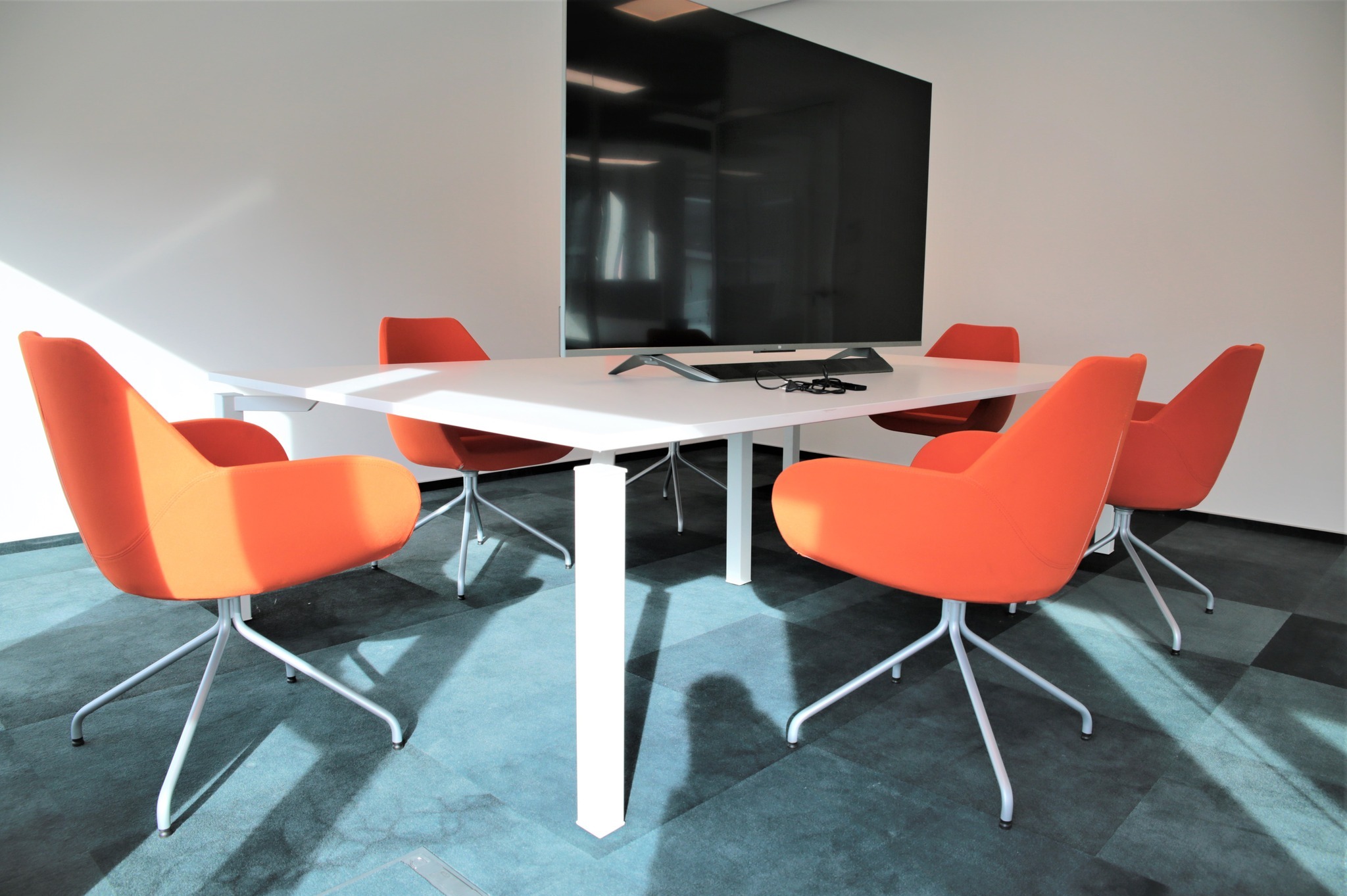 Nowoczesna sala konferencyjna: biały stół z centralną nogą, otoczony pomarańczowymi krzesłami na srebrnych nogach. W tle duży telewizor na ścianie.