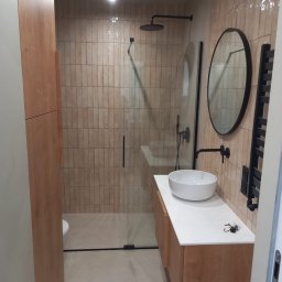 71INSIDE - Nowoczesna łazienka po remoncie: kabina prysznicowa ze szklaną ścianą, umywalka nablatowa na drewnianej szafce, okrągłe lustro i beżowe płytki na ścianach.