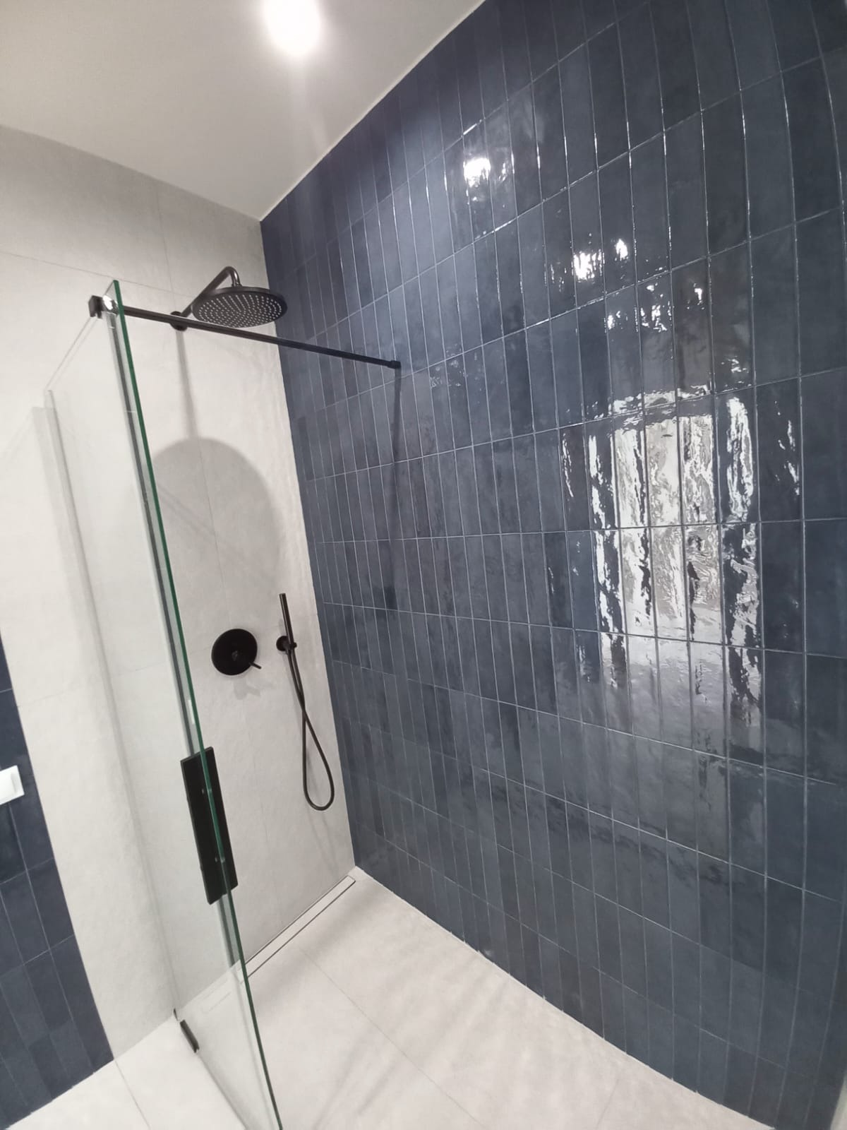 Nowoczesna kabina prysznicowa z grafitowymi, błyszczącymi płytkami ułożonymi pionowo, czarna armatura i szklana ściana. Minimalistyczny design, jasna podłoga.