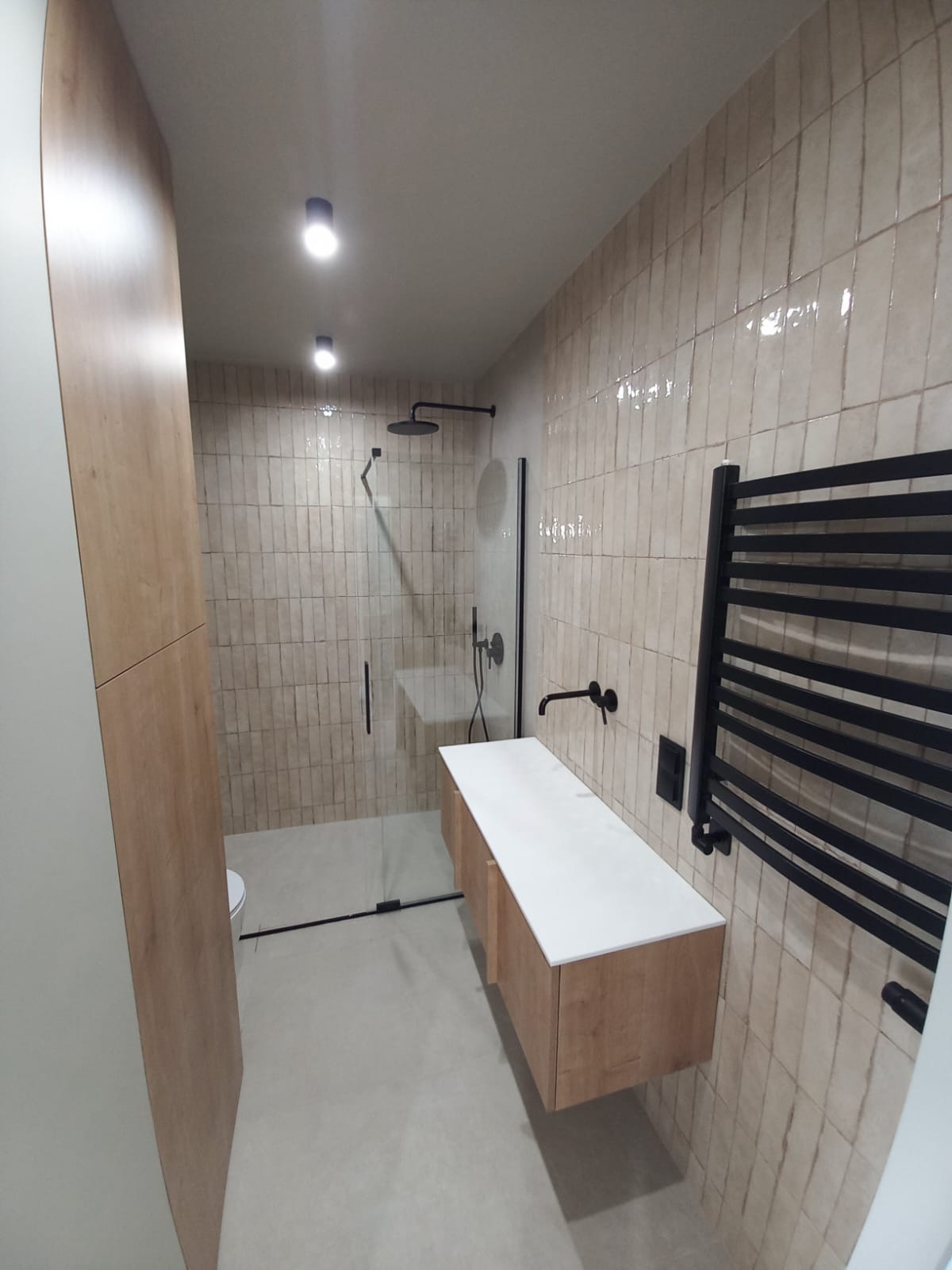 Nowoczesna łazienka z kabiną typu walk-in, beżowymi płytkami imitującymi cegłę, drewnianą szafką pod umywalką i czarnym grzejnikiem drabinkowym.