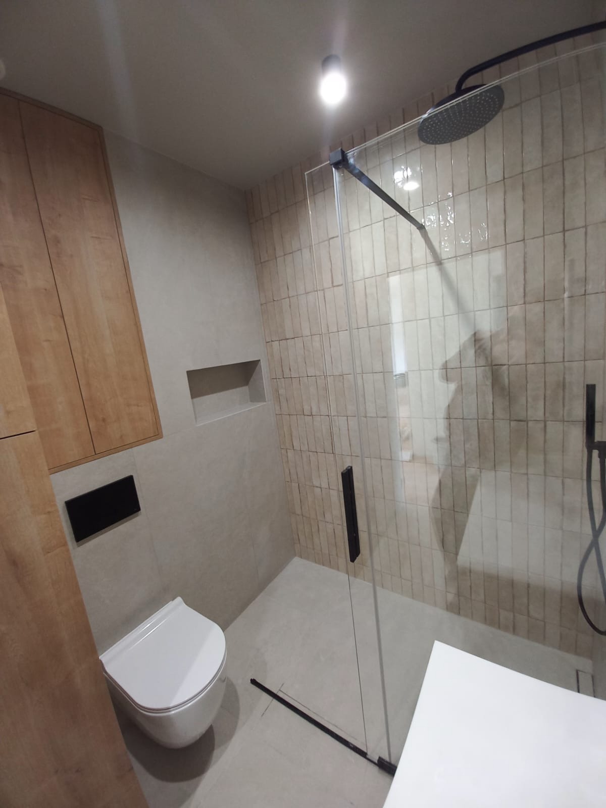 Nowoczesna łazienka z kabiną prysznicową typu walk-in, beżowymi płytkami imitującymi cegłę, podwieszaną toaletą i drewnianą szafką. Minimalistyczny design.