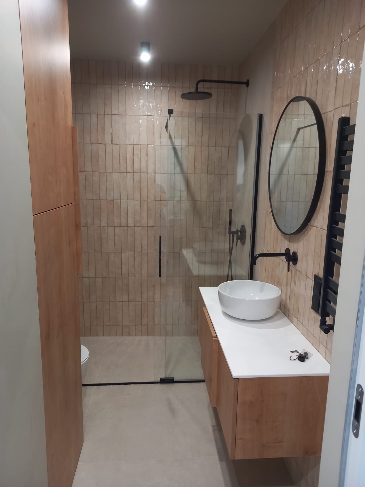 Nowoczesna łazienka po remoncie: kabina prysznicowa ze szklaną ścianą, umywalka nablatowa na drewnianej szafce, okrągłe lustro i beżowe płytki na ścianach.