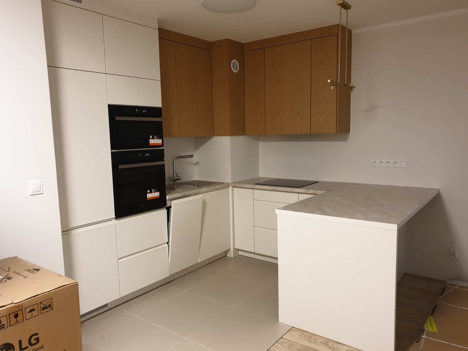 Nowoczesna kuchnia z białymi szafkami i drewnianymi górnymi szafkami. Widoczne sprzęty AGD i oświetlenie. Na podłodze jasne płytki. Częściowo widoczny karton.