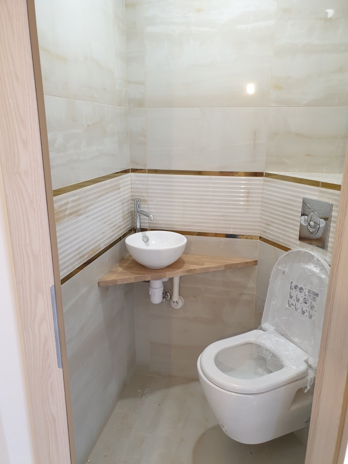 Nowoczesna, mała łazienka po remoncie: biała umywalka na drewnianym blacie narożnym, podwieszana toaleta z folią ochronną, beżowe płytki z dekoracyjnym paskiem.