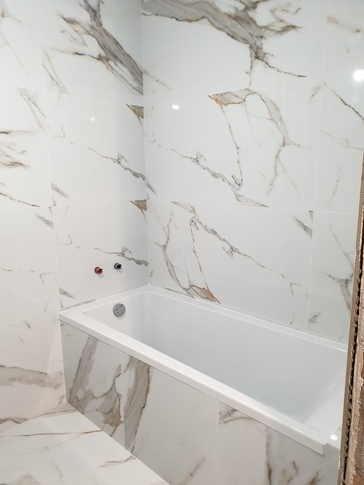 Biała wanna obudowana płytkami imitującymi marmur, z widocznymi zaworami wody. Ściany i podłoga wykończone w tym samym stylu. Minimalistyczny design łazienki.