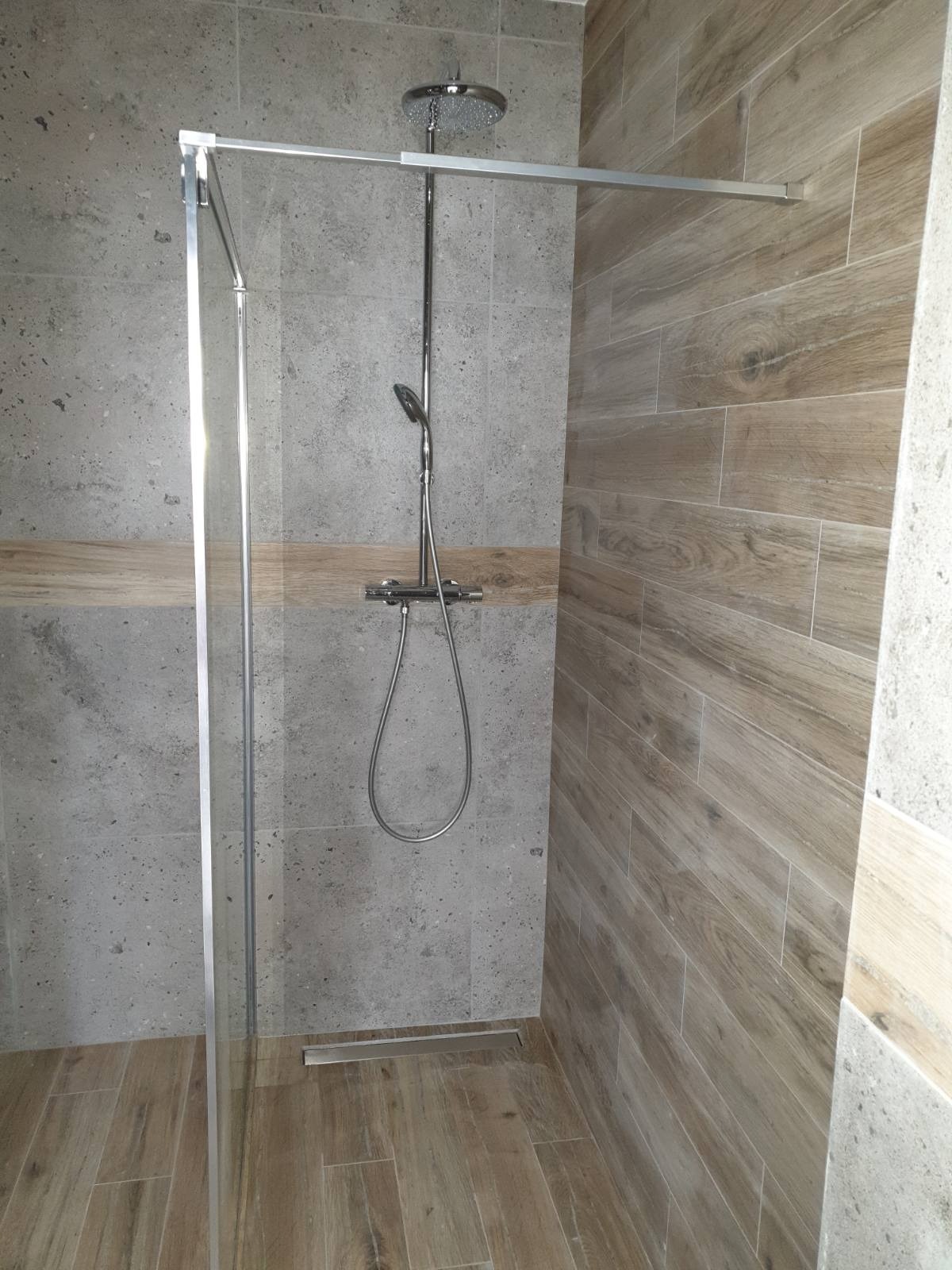 Nowoczesna kabina prysznicowa z szarymi płytkami imitującymi kamień i drewnopodobnymi płytkami. Widoczny odpływ liniowy i chromowana armatura.
