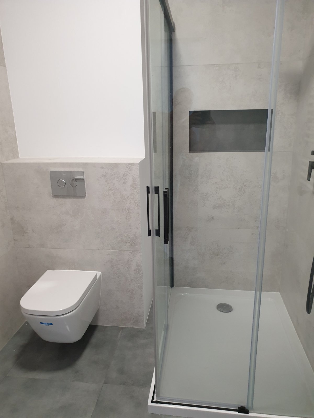 Nowoczesna łazienka po remoncie: kabina prysznicowa z czarnymi detalami, podwieszana toaleta i płytki imitujące kamień. Minimalistyczny design z wnęką w ścianie.