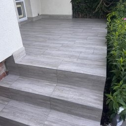 Jakub Szyptur - Trzy stopnie i zadaszony taras wyłożone szarymi, drewnopodobnymi płytkami ceramicznymi w przesuniętym wzorze, z zielenią i białymi ścianami w tle.