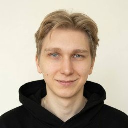 MG GROUP SPÓŁKA Z OGRANICZONĄ ODPOWIEDZIALNOŚCIĄ - Portret młodego mężczyzny o jasnych włosach i niebieskich oczach, ubranego w czarną bluzę z kapturem, na jasnym tle. Widoczny łańcuszek na szyi.