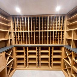 Grzegorz Augustynek - Drewniana zabudowa wnęki z półkami i przegródkami na wino, wykończona blatem z ciemnego kamienia. Oświetlenie sufitowe podkreśla naturalny odcień drewna.