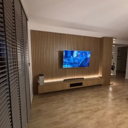 Grzegorz Augustynek - Drewniana zabudowa ściany z telewizorem i podświetleniem LED, zintegrowana z szafką RTV. Na pierwszym planie żaluzje w kolorze szarym.