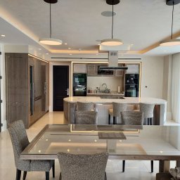 Grzegorz Augustynek - Nowoczesna kuchnia z wyspą i stołem jadalnym. Jasne wnętrze z oświetleniem LED. Krzesła tapicerowane w neutralnych barwach. Duże okno z widokiem na zieleń.