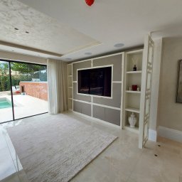 Grzegorz Augustynek - Nowoczesny salon z zabudową TV w odcieniach bieli i szarości, z otwartą przestrzenią na basen. Minimalistyczny design, jasne wnętrze, elegancki dywan.