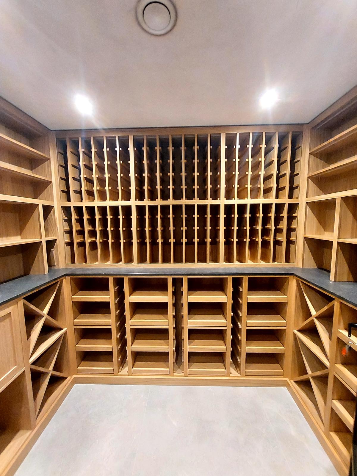 Drewniana zabudowa wnęki z półkami i przegródkami na wino, wykończona blatem z ciemnego kamienia. Oświetlenie sufitowe podkreśla naturalny odcień drewna.