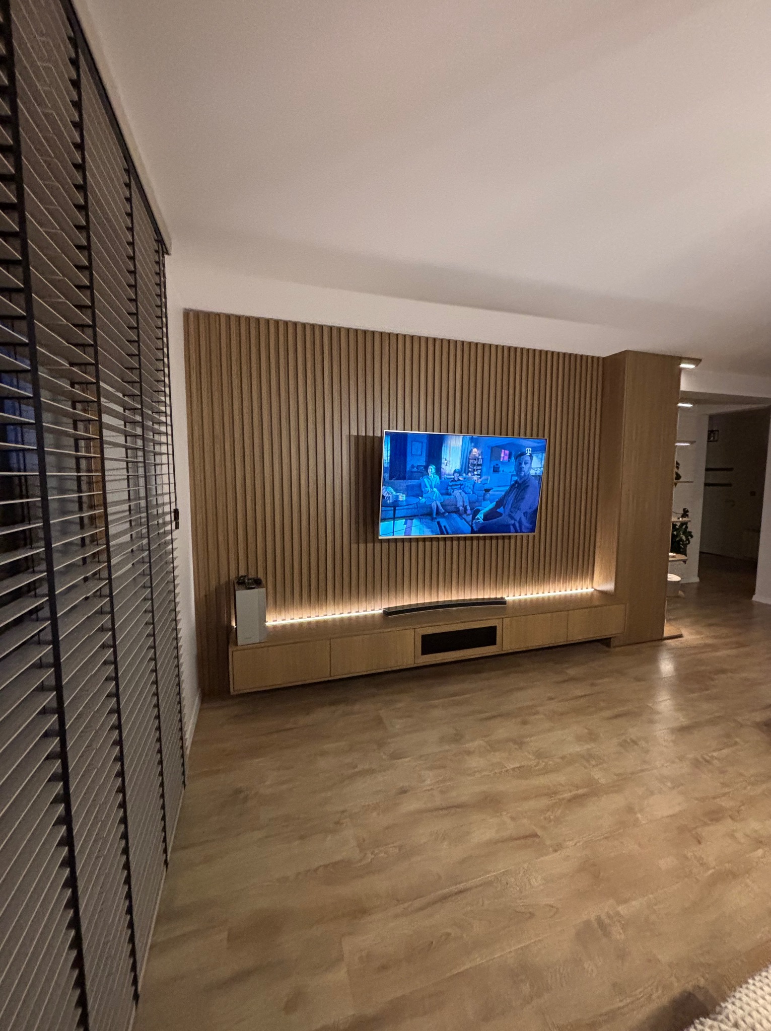 Drewniana zabudowa ściany z telewizorem i podświetleniem LED, zintegrowana z szafką RTV. Na pierwszym planie żaluzje w kolorze szarym.