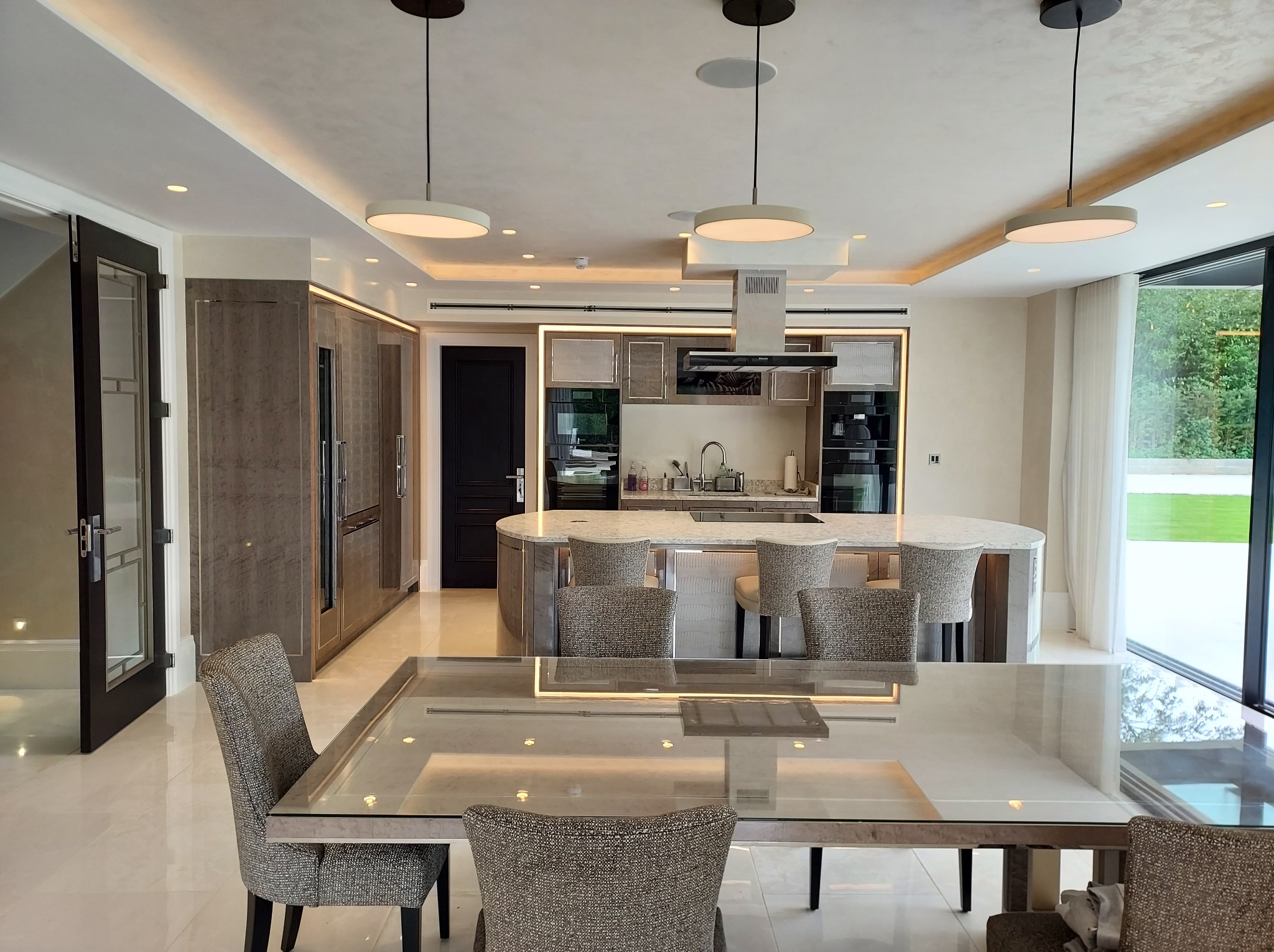 Nowoczesna kuchnia z wyspą i stołem jadalnym. Jasne wnętrze z oświetleniem LED. Krzesła tapicerowane w neutralnych barwach. Duże okno z widokiem na zieleń.