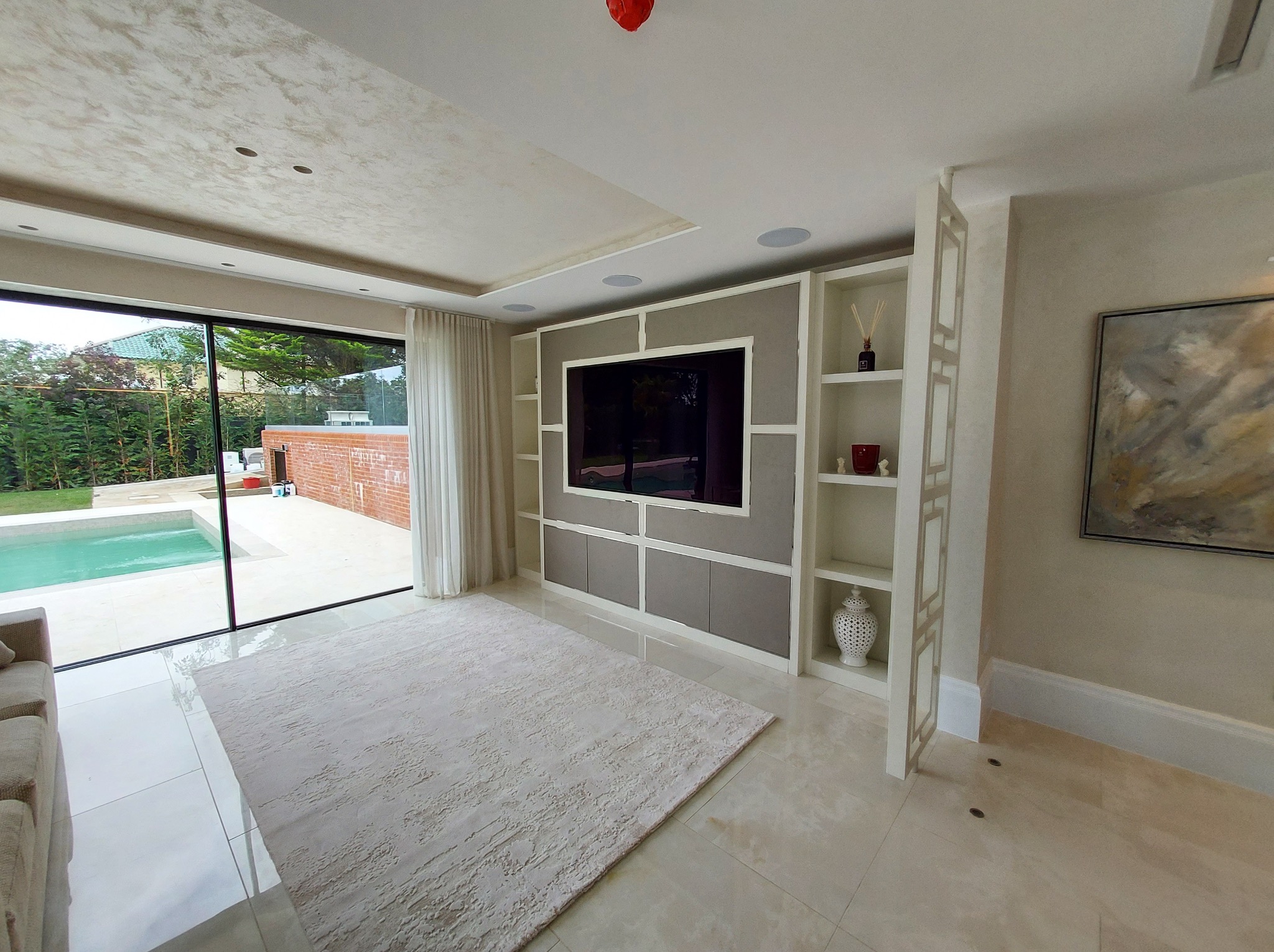 Nowoczesny salon z zabudową TV w odcieniach bieli i szarości, z otwartą przestrzenią na basen. Minimalistyczny design, jasne wnętrze, elegancki dywan.