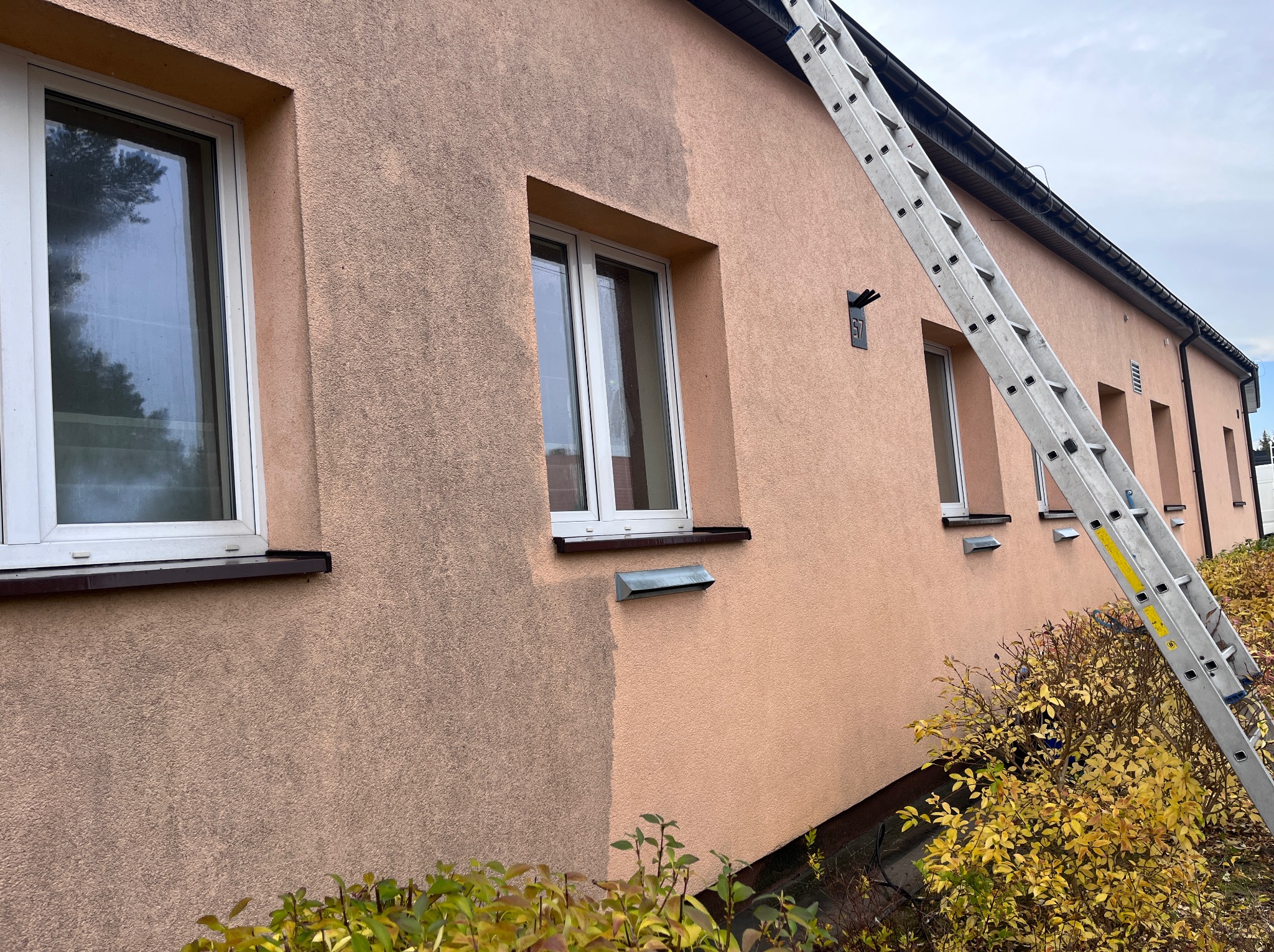 Częściowo umyta, różowa elewacja budynku z oknami. Widoczna drabina oparta o ścianę. Efekt czyszczenia elewacji, kontrast brudnej i czystej powierzchni.